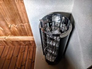 Nærbillede af en cylindrisk elektrisk Harvia saunaovn med stenkurv i rustfrit stål i TimberIN Nordic Oval™ Terrace sauna, ved en træbænk og grå varmeskærm