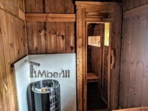 TimberIN Nordic Oval™ Terrace havesauna – forrummet indefra med sort saunaovn foran en beskyttelsesplade i metal, trædør til saunarummet og en lille bænk