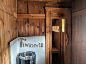 TimberIN Nordic Oval™ Terrace havesauna – blik gennem forrummet med saunaovn og stenkurv foran en beskyttelsesplade i metal samt trædør til bænkerummet