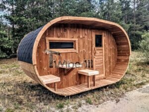 TimberIN Nordic Oval™ Terrace havesauna i trekvartprofil ved skovkanten, med oval træfront, dybt forrum, dør til højre, tværvindue til venstre og udendørs bænke