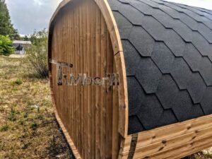 TimberIN Nordic Oval™ Terrace – udendørs sauna med terrasse, med sort tag, gamle træer og en bygning i baggrunden, på en åben eng