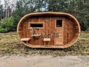 TimberIN Nordic Oval™ Terrace havesauna set lige forfra i skoven, med symmetrisk oval facade, tværvindue til venstre, trædør til højre og tre små bænke