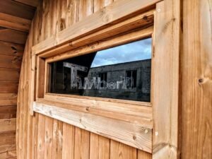 Nærbillede af det vandrette rektangulære sidevindue på TimberIN Nordic Oval™ Terrace sauna, med massiv træramme og omgivelserne spejlet i ruden