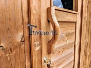Detalje af den lukkede trædør på TimberIN Nordic Oval™ Terrace sauna, med sort metalbeslag, buet træhåndtag og et lille vindue ovenover