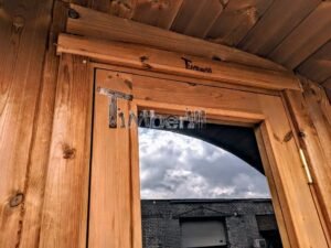 TimberIN Nordic Oval™ Terrace – udendørs sauna med terrasse, med åben dør og en bygning i baggrunden, under åben himmel