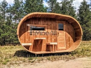 TimberIN Nordic Oval™ Terrace havesauna set forfra i solskin, med oval træramme, tværgående vindue, lukket dør og et opstillet træelement i forrummet