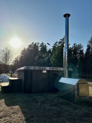 TimberIN Nordic Square™ Acryl – kvadratisk vildmarksbad i akryl, med skorsten i rustfrit stål fra brændeovnen og gamle træer i baggrunden, i en anlagt have
