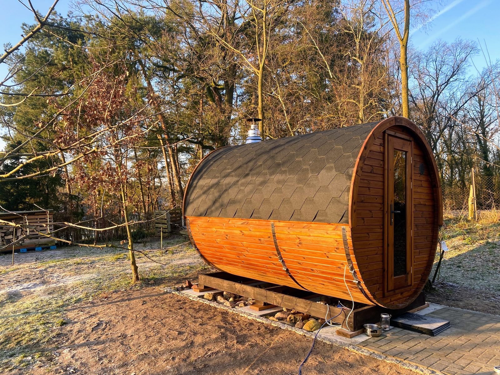 Lille sauna udendørs til 2–4 personer 1 Lille tøndesauna i træ med sort tag og glasdør, placeret på et fundament under træerne i haven