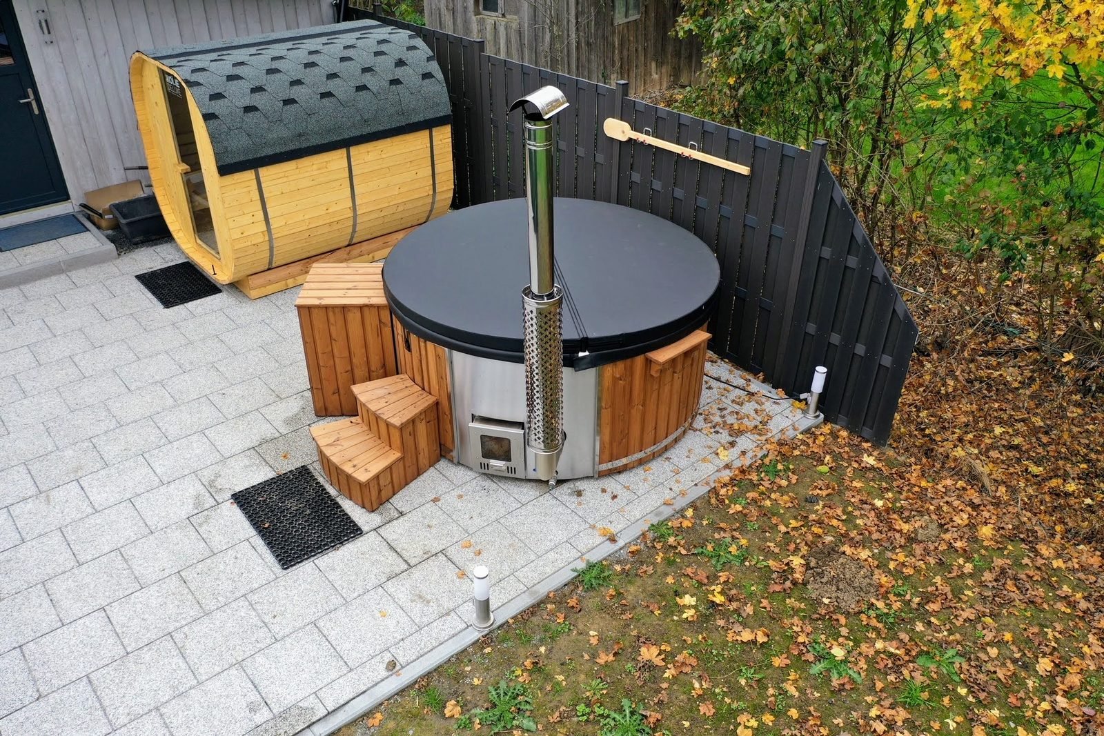 Tøndesauna med glasende og vildmarksbad med låg, trappe og skorsten side om side på en flisebelagt terrasse