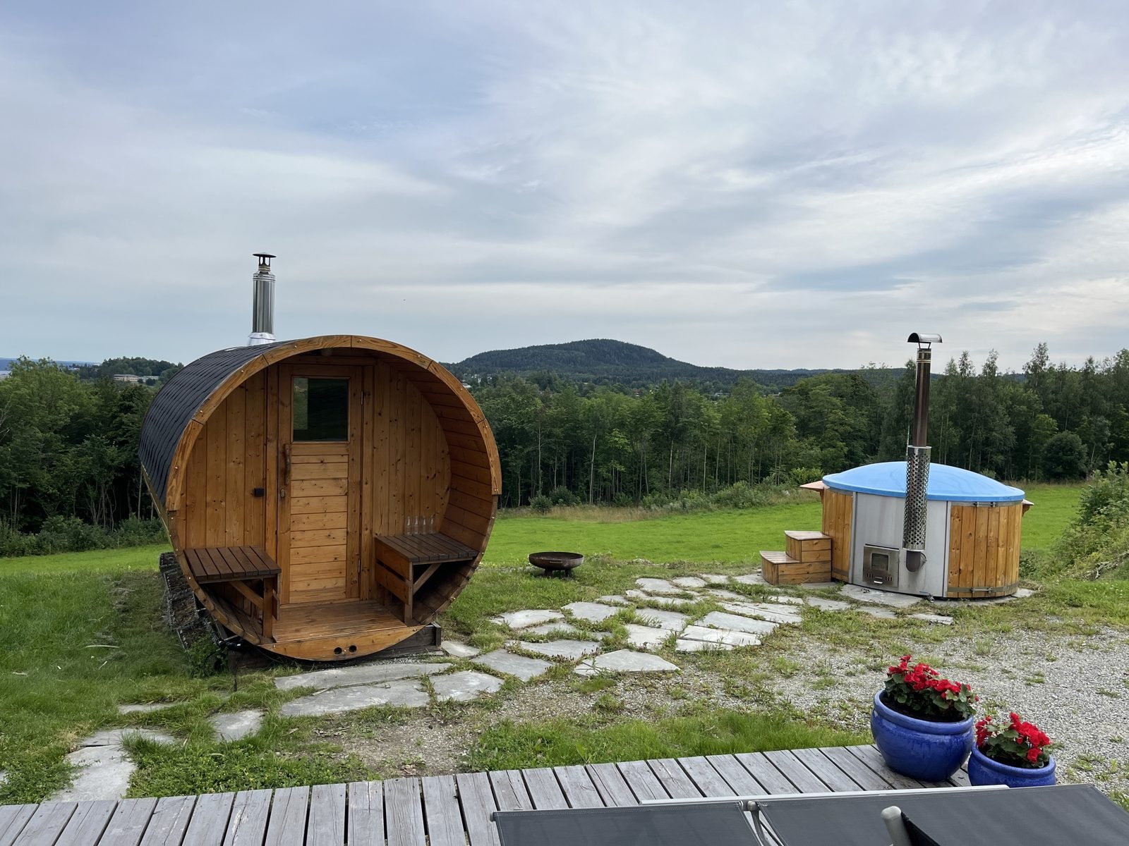 Udendørs sauna med brændeovn 1 Saunatønde i træ med skorsten fra brændeovnen og terrasse foran døren på en græsmark
