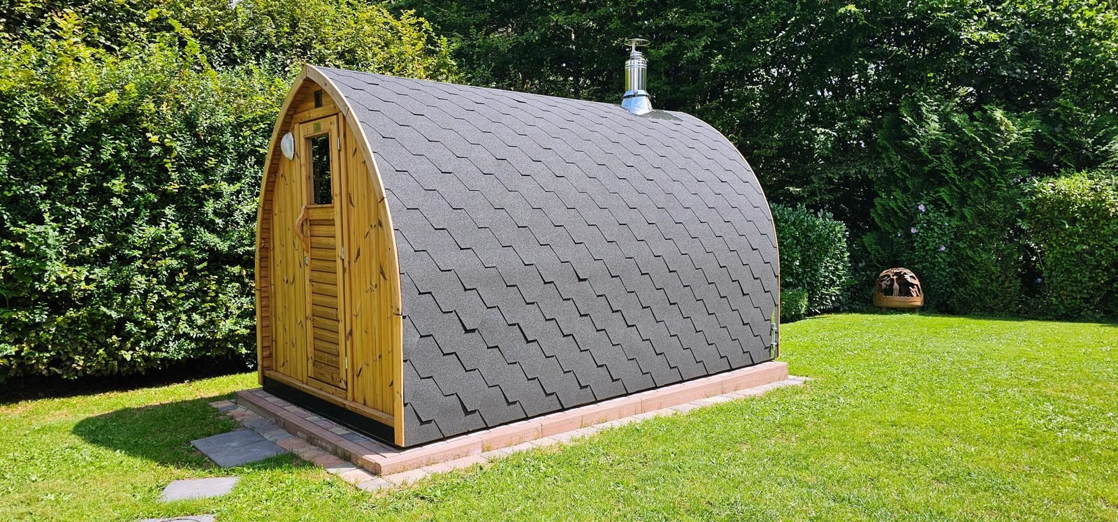 Udendørs pod-sauna i træ med tagshingles på en græsplæne i haven
