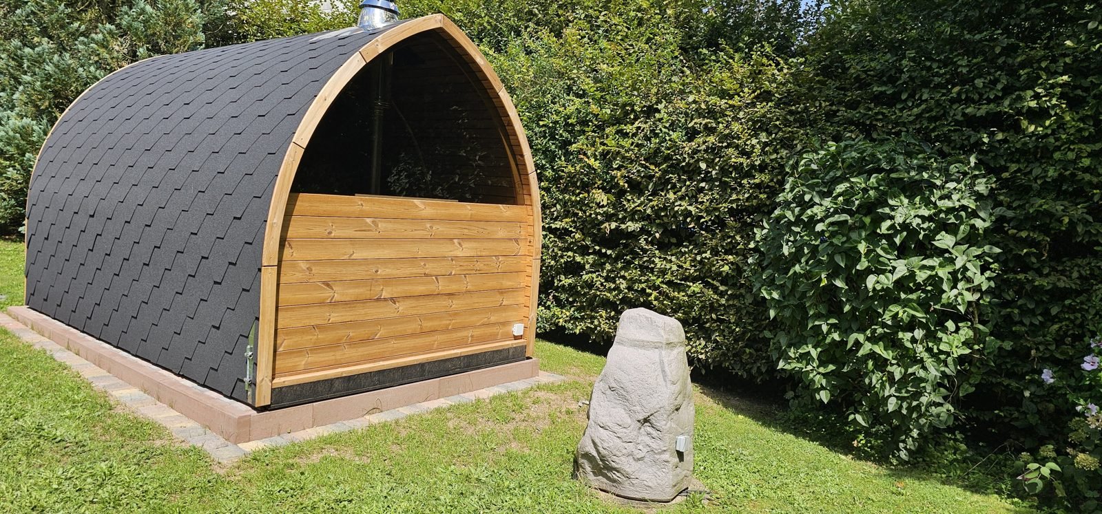Udendørs pod-sauna i træ med sorte tagshingles på et fundament i en grøn have