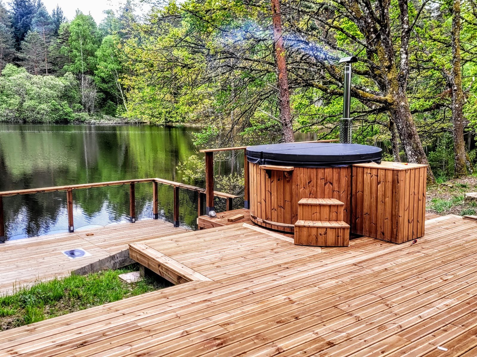 TimberIN® Danmark 1 Rundt vildmarksbad i træ med låg og trappe på en træterrasse ved en skovsø