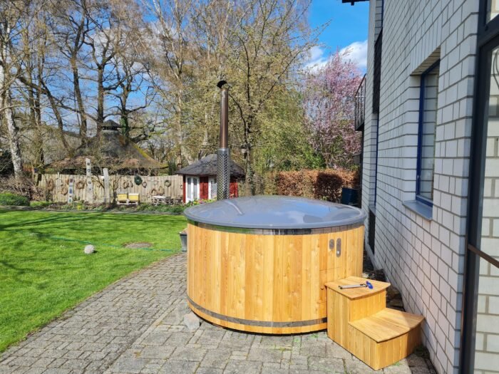 Vildmarksbad i træ med låg placeret på jævn flisebelægning op ad husmuren