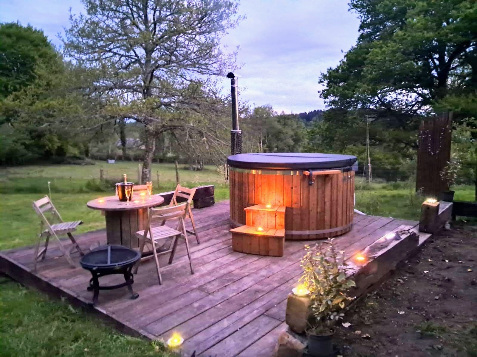 Vildmarksbad 1 Vildmarksbad i træ med varmt lys på en træterrasse i haven ved skumringstid, med bord og stole